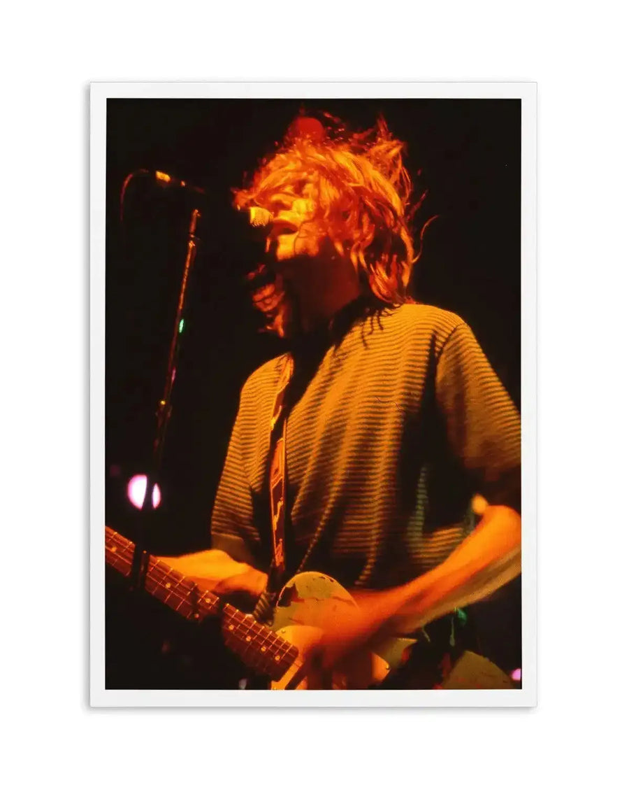 "Photography art print featuring a close-up of Kurt Cobain performing live on stage, bathed in warm orange stage lights. His long, messy blonde hair is thrown back, and he's singing into a microphone while playing a light-colored electric guitar. He wears a horizontally striped shirt, and the image captures the raw energy of a live performance with blurred motion in his hands and hair."