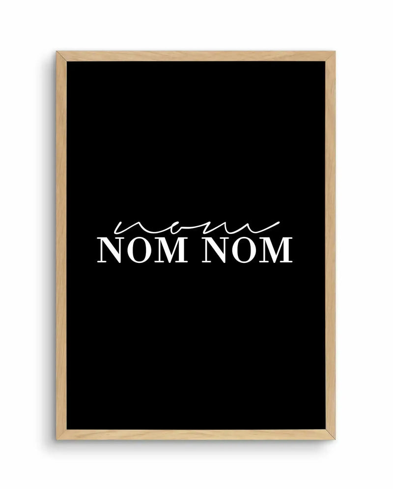 Art print with natural wood frame featuring black background and white text reading "nom nom nom" in a stylized script font above the word "NOM NOM" in bold capital letters. The design uses a minimalist aesthetic with high contrast between the black and white elements.