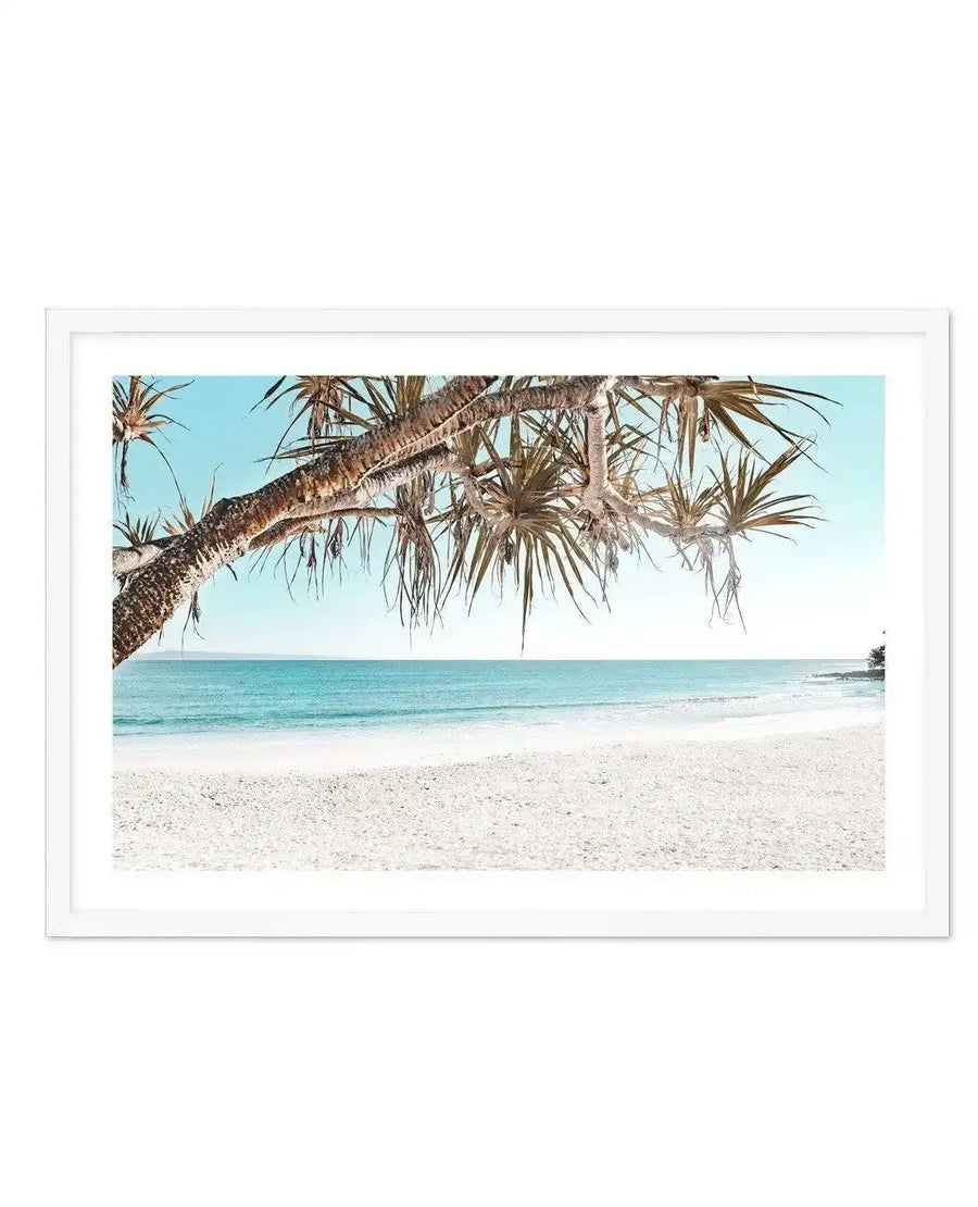 Framed photography art print of a bright coastal beach scene with a white frame and white border. The composition features a white sand beach in the foreground, leading to turquoise ocean waves under a clear light blue sky. A large tree branch with long, spiky green leaves hangs down from the top left, partially obscuring the sky.
