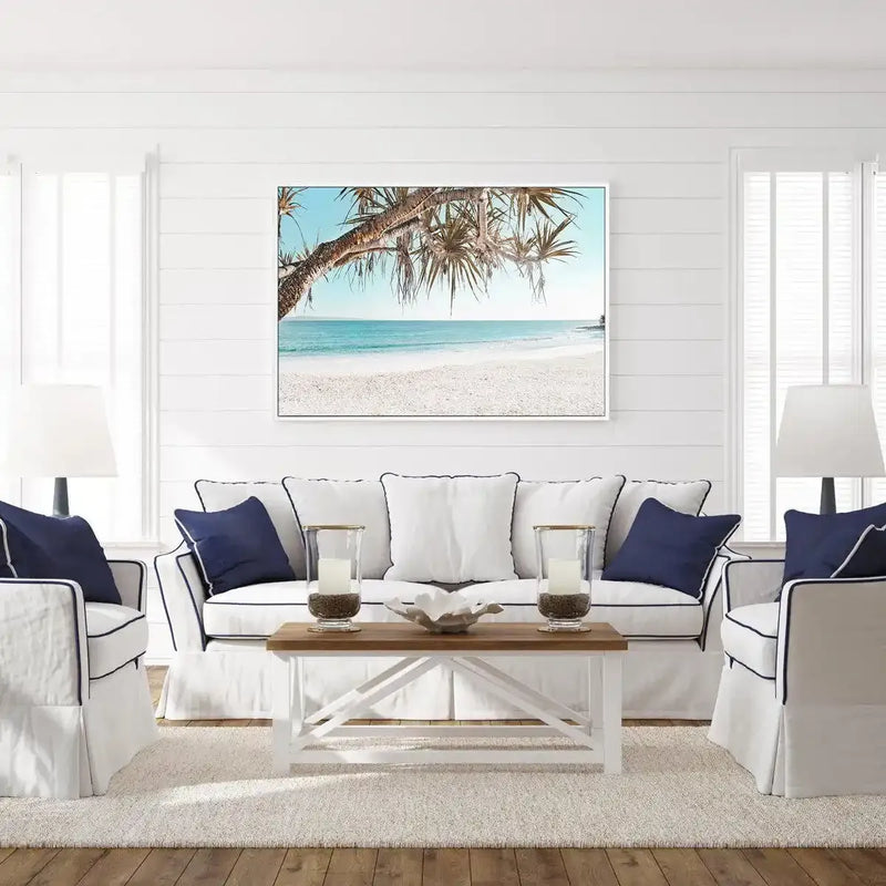 Noosa Days LS framed canvas art print displayed on a white shiplap wall above a cream upholstered sofa with navy blue pillows. The artwork features a tropical beach scene photographed from beneath a palm tree, showing turquoise water, white sand, and clear blue sky, with a black frame and white border. The print hangs centrally in a bright, coastal-themed living room with white furnishings, wooden coffee table, and large windows.
