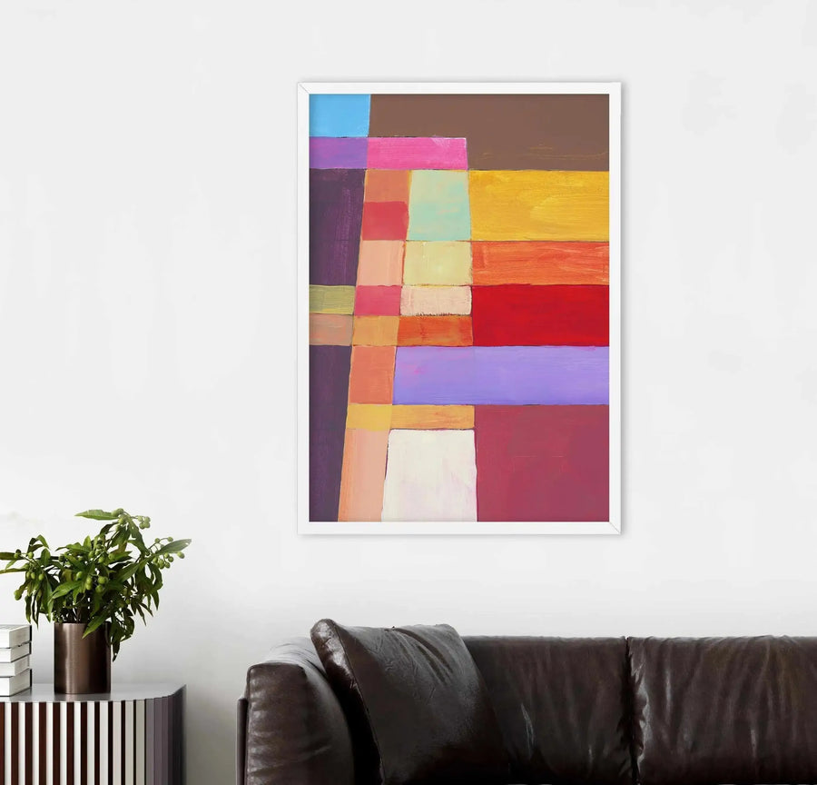 Lifestyle shot of a framed abstract art print displayed above a dark brown leather sofa in a modern living room. The print features a grid-like composition of various sized rectangular blocks of colour, including bright blue, deep purple, hot pink, warm yellow, orange, red, and light lavender. The room also includes a dark brown cylindrical planter with a green leafy plant and a striped cabinet.