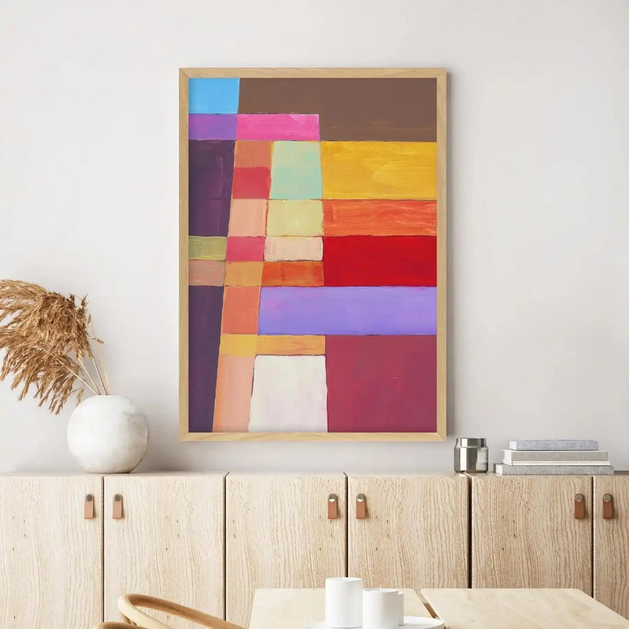 Large framed abstract art print displayed above a light wood console table in a minimalist living room. The print features a grid-like composition of various sized rectangular blocks in a vibrant palette of blue, purple, pink, brown, yellow, orange, red, and cream, with a natural wood frame. The room includes a white ceramic vase with dried pampas grass, a silver canister, and a stack of books on the console table.