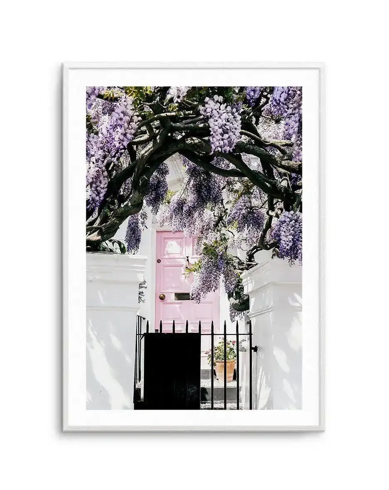 Framed photography art print featuring a charming pink door framed by a vibrant arch of purple wisteria, with a white frame and white border. The composition captures the lush purple blossoms cascading over a dark, gnarled vine, with a black metal gate and white pillars in the foreground.