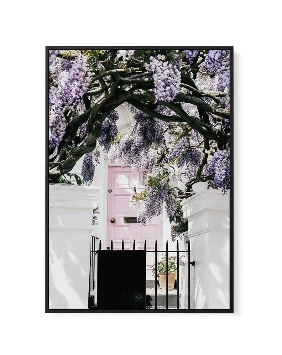 Framed canvas art print featuring a photograph of a pale pink front door on a white townhouse, framed by an ornate dark wooden wisteria vine with cascading lavender and purple flowers overhead. Black wrought iron fence visible at street level. White mat border surrounding the image. Black frame.