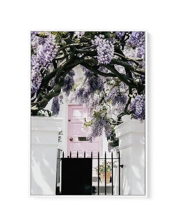 Framed canvas art print featuring a photograph of a white townhouse with a pale pink front door beneath an ornate black iron gate, all framed by cascading wisteria with lavender-purple flowers and dark twisted branches. The composition captures the romantic charm of a Notting Hill London street facade. The canvas is mounted in a white frame with white border.