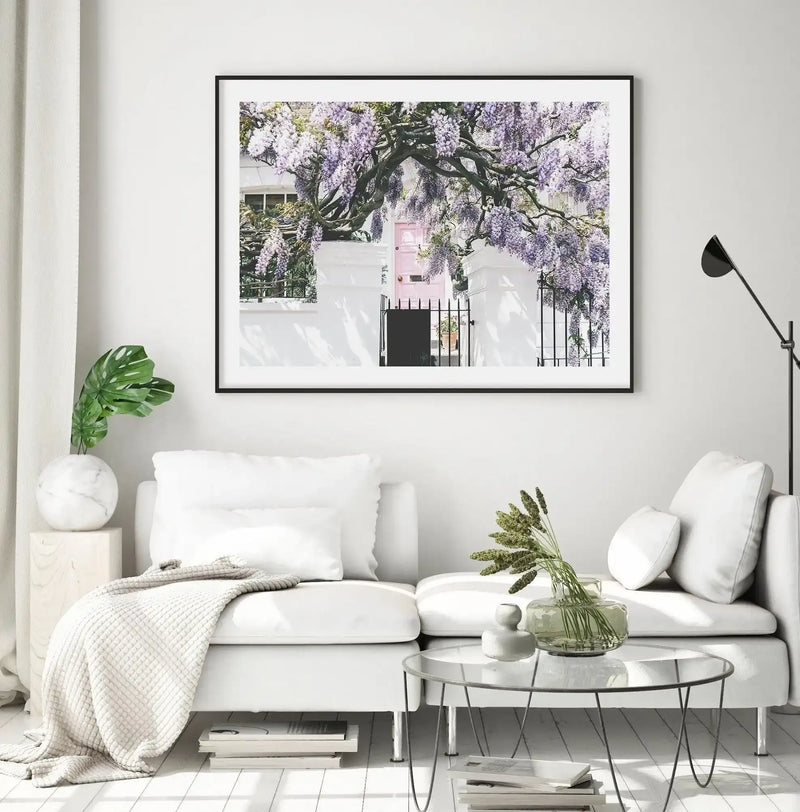 Large framed art print displayed above a white sectional sofa in a bright, minimalist living room. The print is a photograph of a wisteria-covered house in Notting Hill, London, featuring a pale pink door surrounded by lush purple wisteria blooms and dark iron fencing, with a black frame and white border. The room includes a white textured throw, a monstera plant in a marble vase, and a clear glass coffee table with dried grasses.