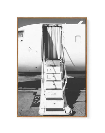Black and white framed canvas art print depicting a close-up view of an airplane stairway with metal steps and white railings extending downward. The fuselage of a white commercial aircraft is visible in the background with an open doorway and cream-colored curtains. The composition captures the geometric lines of the stairs against the tarmac and aircraft body, creating strong contrast and depth. The artwork is presented in a natural wood-tone frame with clean lines, mounted on a white border.