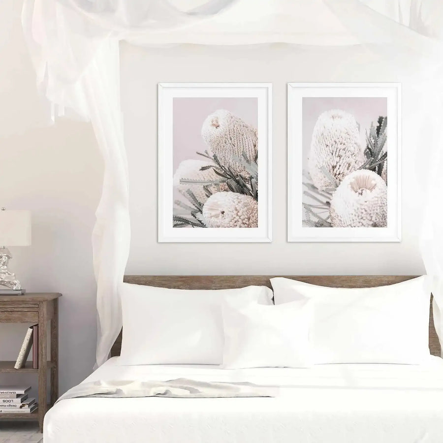 Two framed art prints hung above a wooden bed headboard in a minimalist bedroom. Each print features close-up photography of delicate banksia flowers with fuzzy, cream-colored blooms and dark green stems against soft pink and gray backgrounds. The prints are in white frames with white borders. The bedroom setting includes white bedding, a rustic wooden nightstand with a decorative lamp, sheer white curtains, and light neutral walls, creating a calm, botanical-themed aesthetic.
