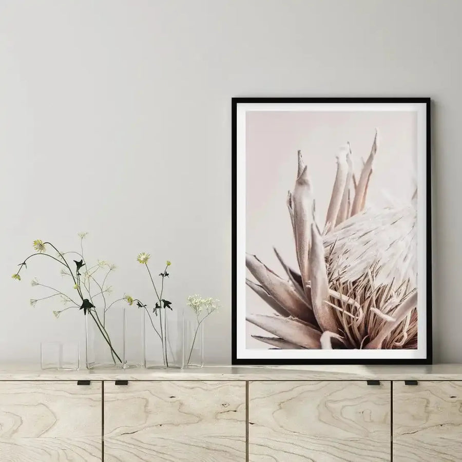 Framed art print displayed on a light wood dresser featuring a close-up photograph of a beige protea flower with soft, muted tones. The protea's layered petals and spiky leaves are rendered in dusty tan, cream, and warm brown hues against a pale background. The print is mounted in a black frame with a white border. To the left of the frame sit several clear glass bud vases containing delicate green stems and small white flowers, styled against a neutral wall.