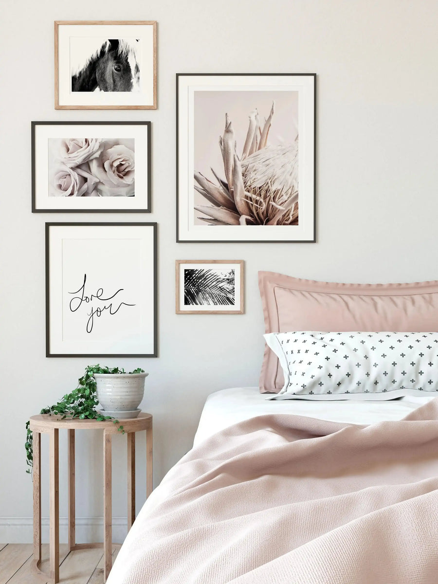 Beige Protea art print displayed in a natural wood frame with white border, hanging above a wooden bedside table with potted plant. The print features a close-up photograph of a protea flower with soft taupe and cream tones, showcasing layered petals and delicate texture against a neutral beige background. Part of a gallery wall arrangement above a bed with blush pink and patterned bedding.