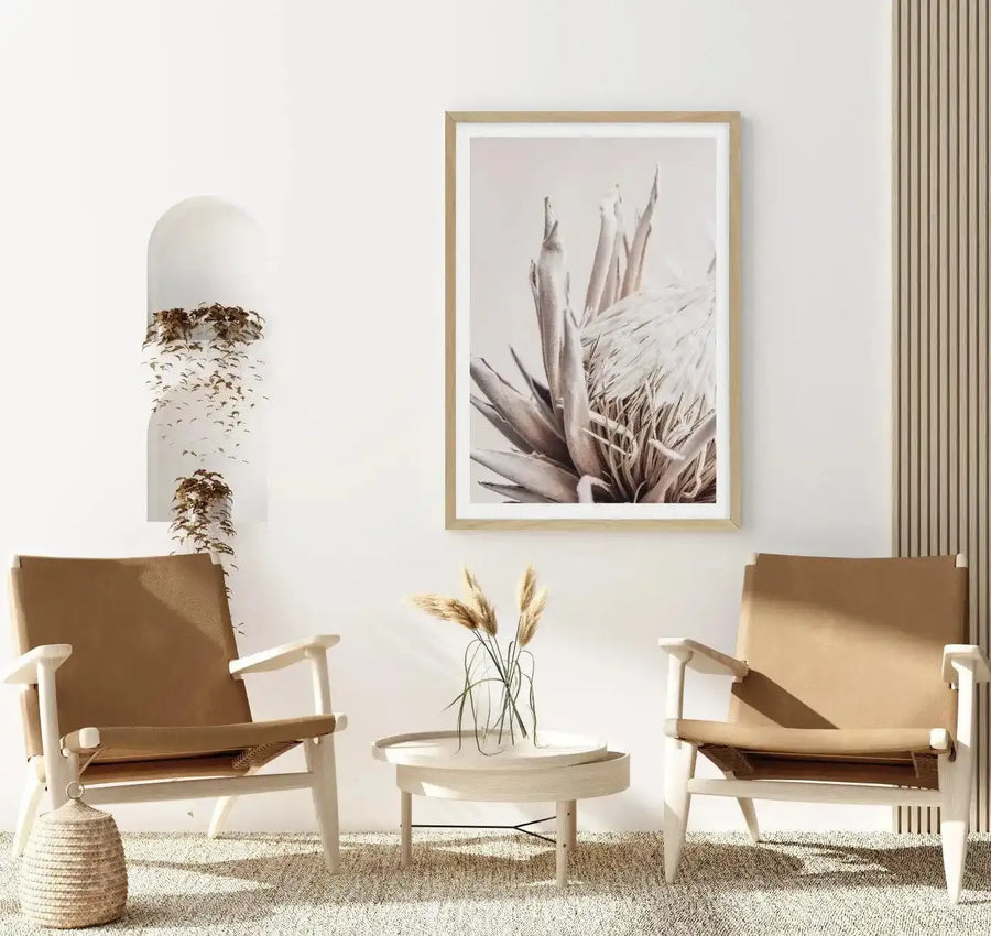 Beige Protea art print in natural wood frame displayed on white wall above two cream canvas chairs flanking small round wooden table with dried grasses in glass vase. Print shows close-up botanical illustration of protea flower with soft taupe and gray tones, delicate pencil-like detailing on petals and leaves, mounted with white border. Neutral interior scene with woven basket, striped curtains, and light-colored area rug.