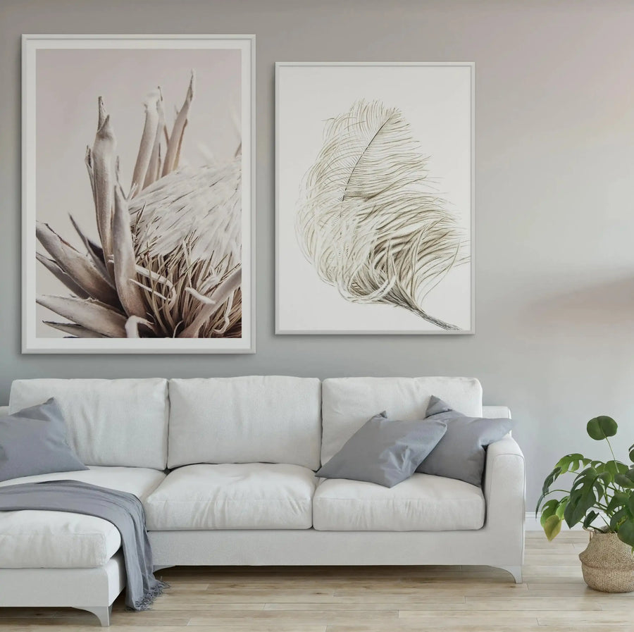 Framed art print featuring a close-up photograph of a beige protea flower with soft brown tones and layered petals, displayed in a white frame with white border above a cream linen sofa in a minimalist living room. Beside it hangs a botanical line drawing of a feathered leaf in muted green and tan tones, also in a white frame. A potted plant with green foliage sits on the floor to the right.