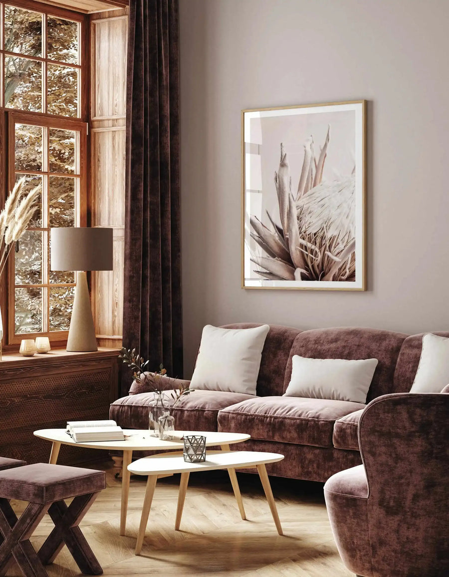 Beige Protea art print displayed in a natural wood frame with white border, hung on a pale taupe wall above a burgundy sofa in a modern living room. The print features a detailed botanical illustration of protea flowers in soft sepia and neutral tones, depicting layered petals and stems in a minimalist style. The framed artwork measures approximately 24 by 36 inches and complements the warm wood tones of the window frame, side table, and natural light flooding the contemporary space.