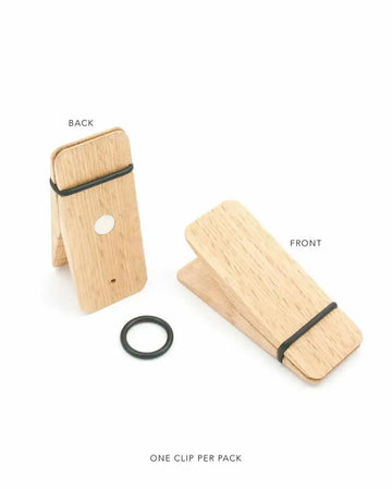 Wooden clip art print displayed in two views. Back view shows a tall, light natural oak wood clip with a dark elastic band and small circular hole detail, standing upright on a kickstand. Front view shows a wider, angled oak wood clip with matching dark elastic band. A dark rubber ring sits separately below. Both pieces feature light wood grain texture and rounded edges, with natural wood finish.