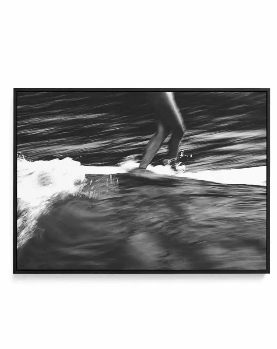 Framed black and white canvas print of a surfer riding a wave, captured with a blurred motion effect. The composition focuses on the lower body of the surfer and the surfboard, with turbulent water and white foam in the foreground and dark, rippling water in the background. The print is displayed in a black frame.