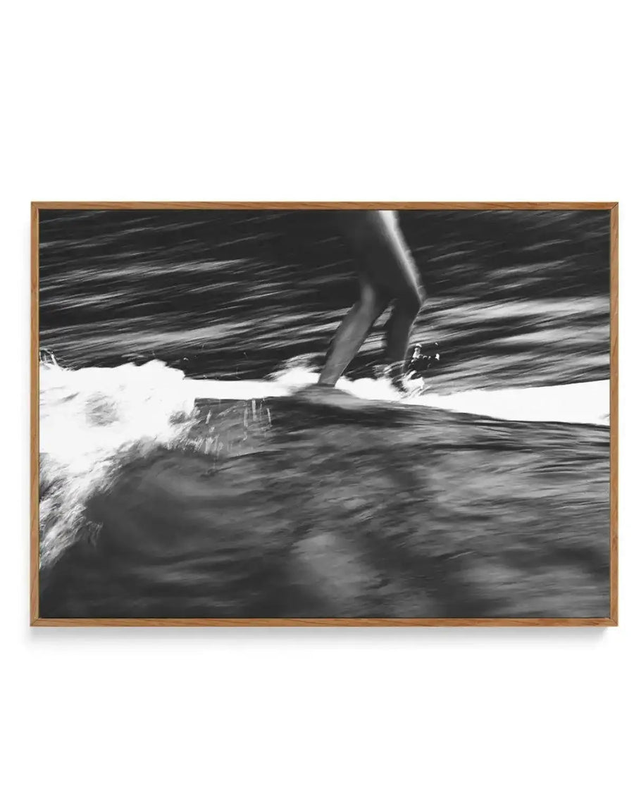 Framed black and white canvas print of a surfer riding a wave, captured with a blurred motion effect. The composition features the lower half of a person's body, legs bent, standing on a surfboard as it cuts through a breaking wave, creating white foam. The background shows dark, blurred water, suggesting speed and movement. The print is displayed in a natural oak frame.