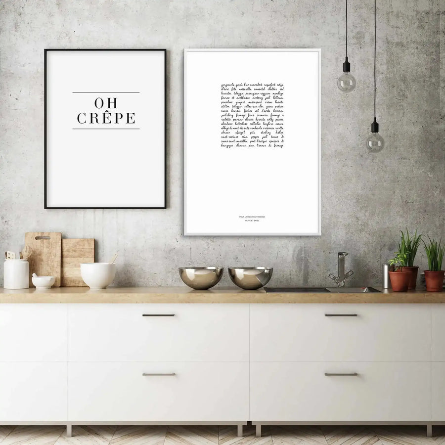 Lifestyle shot of a modern kitchen featuring two framed art prints on a concrete wall above a light wood countertop. The left print, in a black frame with a white border, reads "OH CRÊPE" in black sans-serif font. The right print, in a white frame with a white border, displays a handwritten list of cheese names in black script. The kitchen includes white cabinets, stainless steel bowls, a sink, and potted plants.