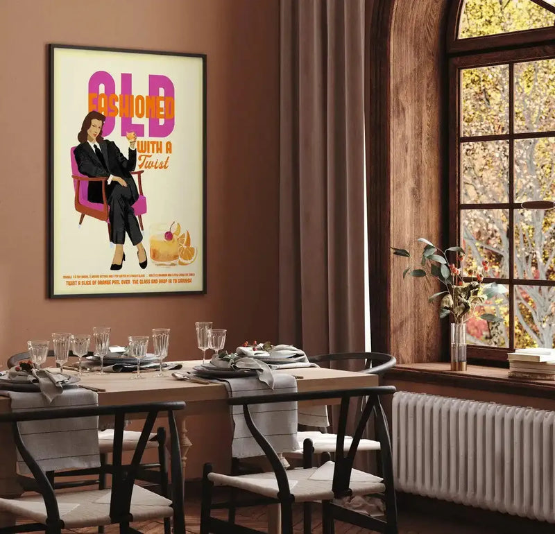 Lifestyle shot of a dining room with an art print displayed on a warm brown wall. The print features a woman in a black pantsuit seated in a pink armchair, holding an orange cocktail. The text "OLD FASHIONED WITH A Twist" is above her. Below the image is text describing how to make the drink. The dining room has a wooden table set with glasses and plates, black chairs, and a large window with wooden framing looking out onto autumn trees. A white radiator is visible below the window.
