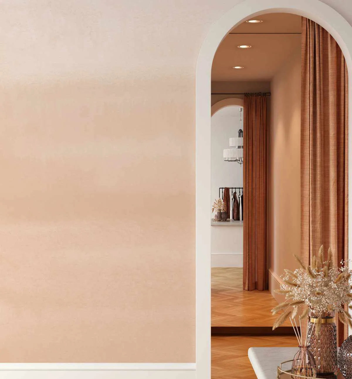 Lifestyle shot of a room with Ombre Soft Terracotta wallpaper applied to the left wall. The wallpaper features a gradient from a light, creamy beige at the top to a soft, warm terracotta orange at the bottom, with a subtle, textured finish. The wall is framed by a white baseboard. To the right, a white arched doorway leads into another room with a warm, inviting atmosphere, featuring a rich terracotta curtain and a parquet wood floor.