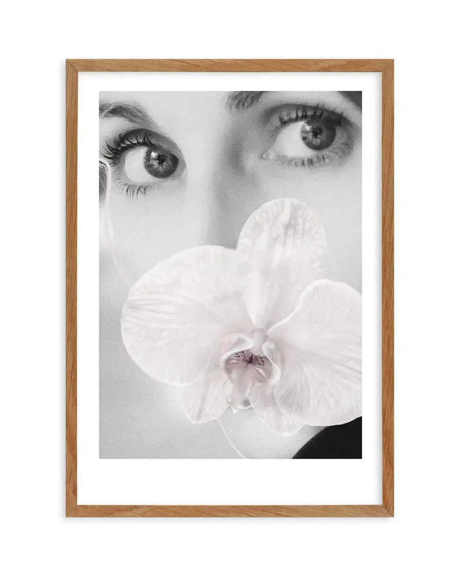 Framed black and white photograph of a woman's face with a white orchid covering her mouth, displayed in a natural oak frame with white border. The woman's eyes are looking upwards and to the left, with long eyelashes visible on her left eye. The orchid has delicate petals and a subtle purple-pink center.