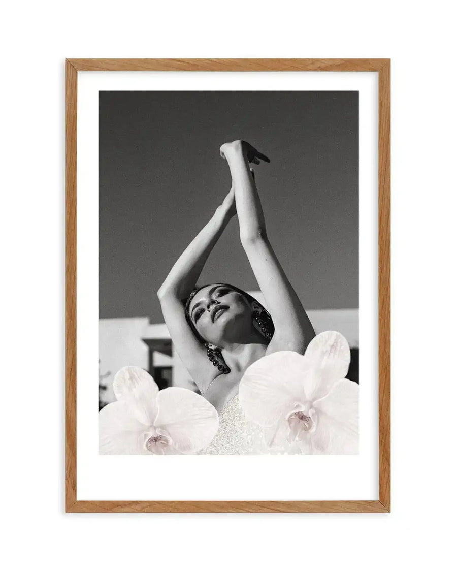 Framed black and white photograph of a woman with her arms raised above her head, looking upwards with a serene expression, displayed in a natural oak frame with white border. The woman is wearing a light-coloured, possibly sequined, top, and large dark earrings. Two large white orchid flowers are superimposed in the foreground, partially obscuring her body. The background is a bright, clear sky and a hint of a building.