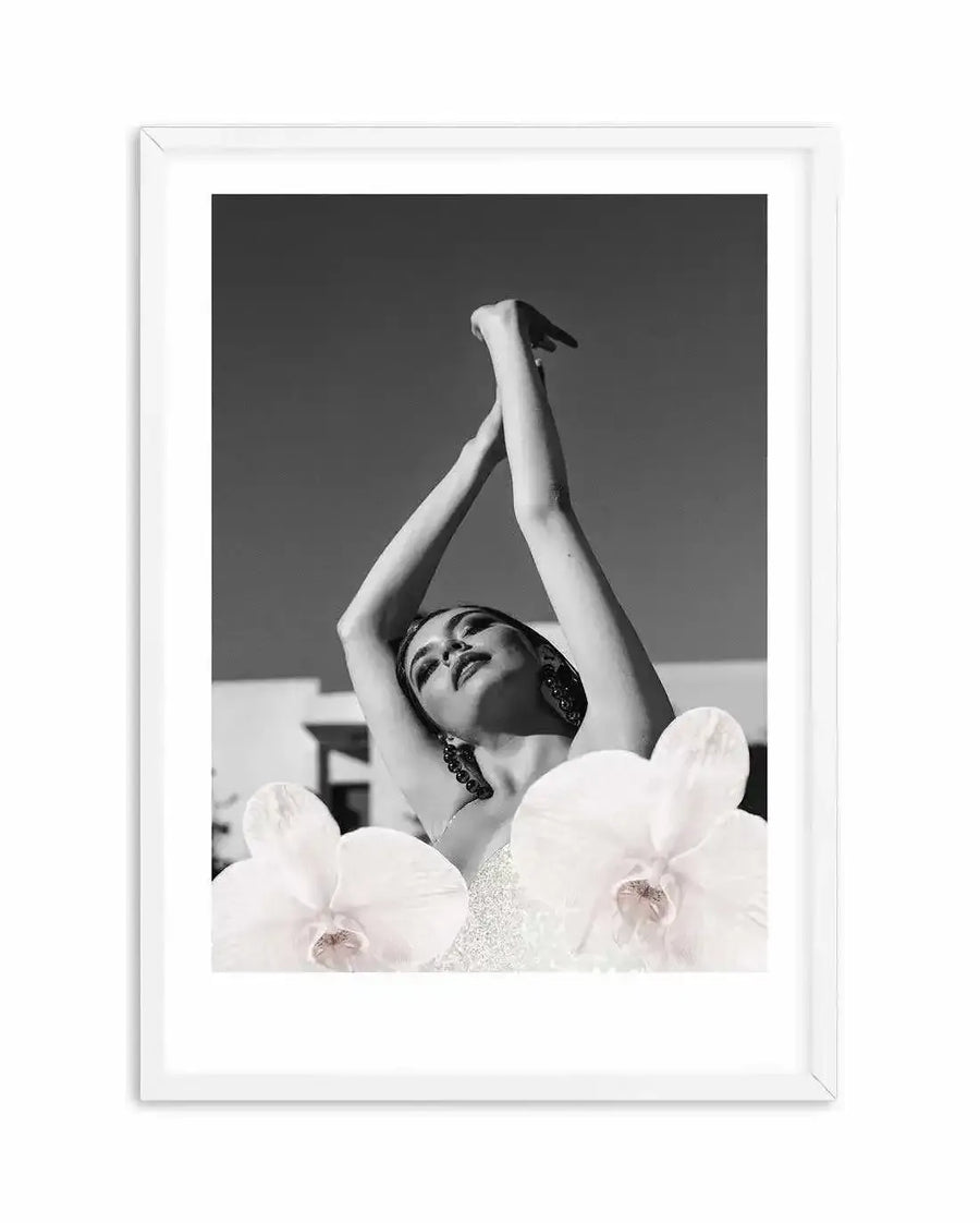 Framed black and white photograph of a woman with her arms raised above her head, looking upwards, with two large white orchids in the foreground, displayed in a white frame with white border. The woman is wearing large earrings and a sparkly top, with a bright sky and building in the background.