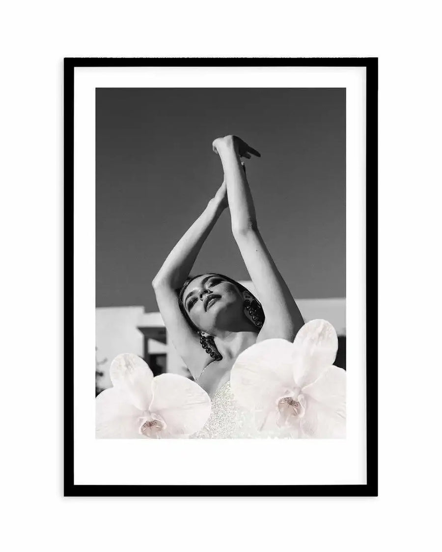 Framed black and white photograph of a woman with her arms raised above her head, looking upwards, with two large white orchid blooms in the foreground. The woman has dark hair and large earrings, and is wearing a light-coloured, possibly sequined, top. The background is a clear sky with a hint of a building structure. The print is displayed in a black frame with a white border.