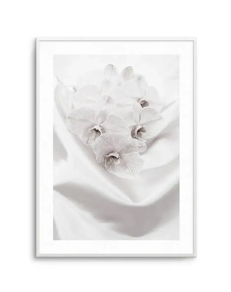Framed art print featuring a delicate white orchid flower cluster with multiple blooms arranged in a shallow white bowl or vessel. The photograph captures fine botanical detail with soft, creamy white petals and subtle brown-grey centers visible on the lower flowers. A soft shadow falls across the composition, creating gentle dimensionality. The image has a serene, minimalist aesthetic with pale neutral tones throughout. The print is presented in a silver-grey frame with a white border.