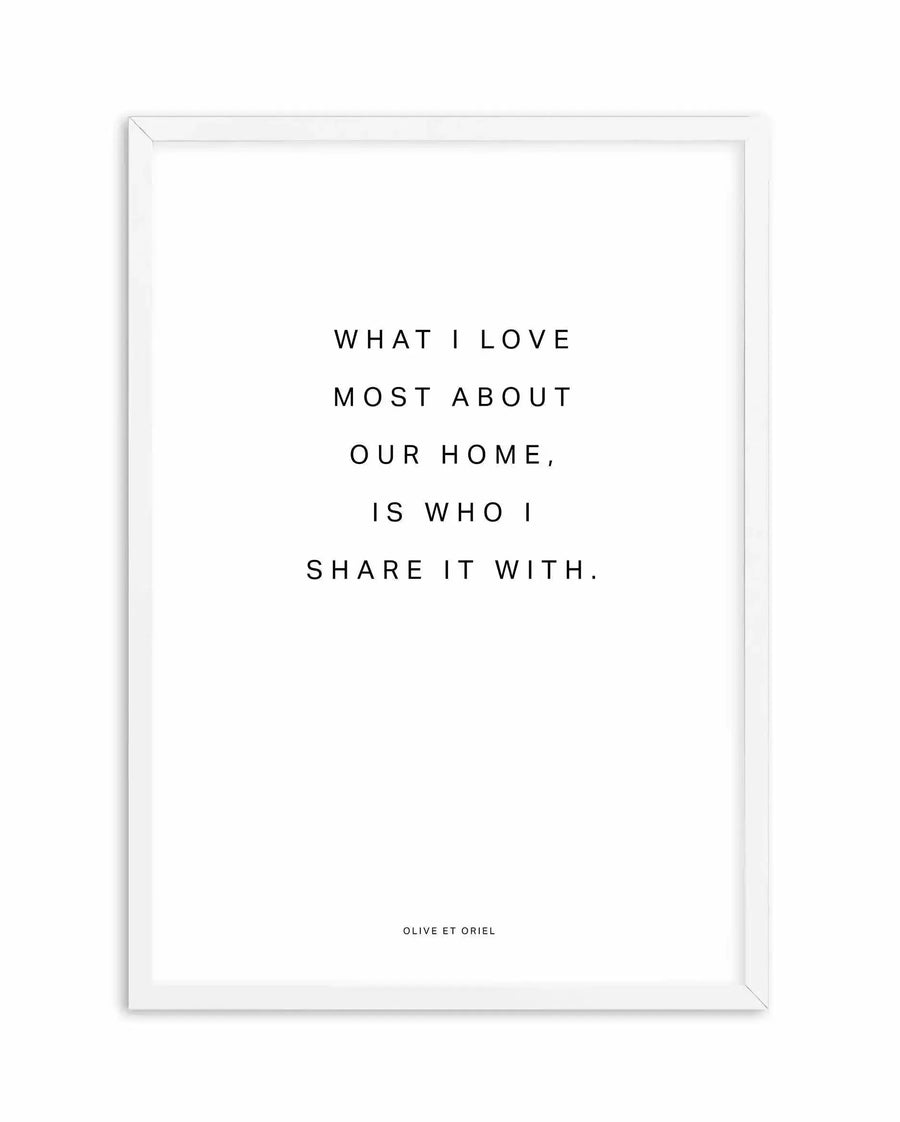 White framed art print featuring centered black text on a white background reading "WHAT I LOVE MOST ABOUT OUR HOME, IS WHO I SHARE IT WITH." with small attribution text at bottom. Clean, minimalist design with sans-serif typography. White wooden frame with subtle 3D depth visible at edges.