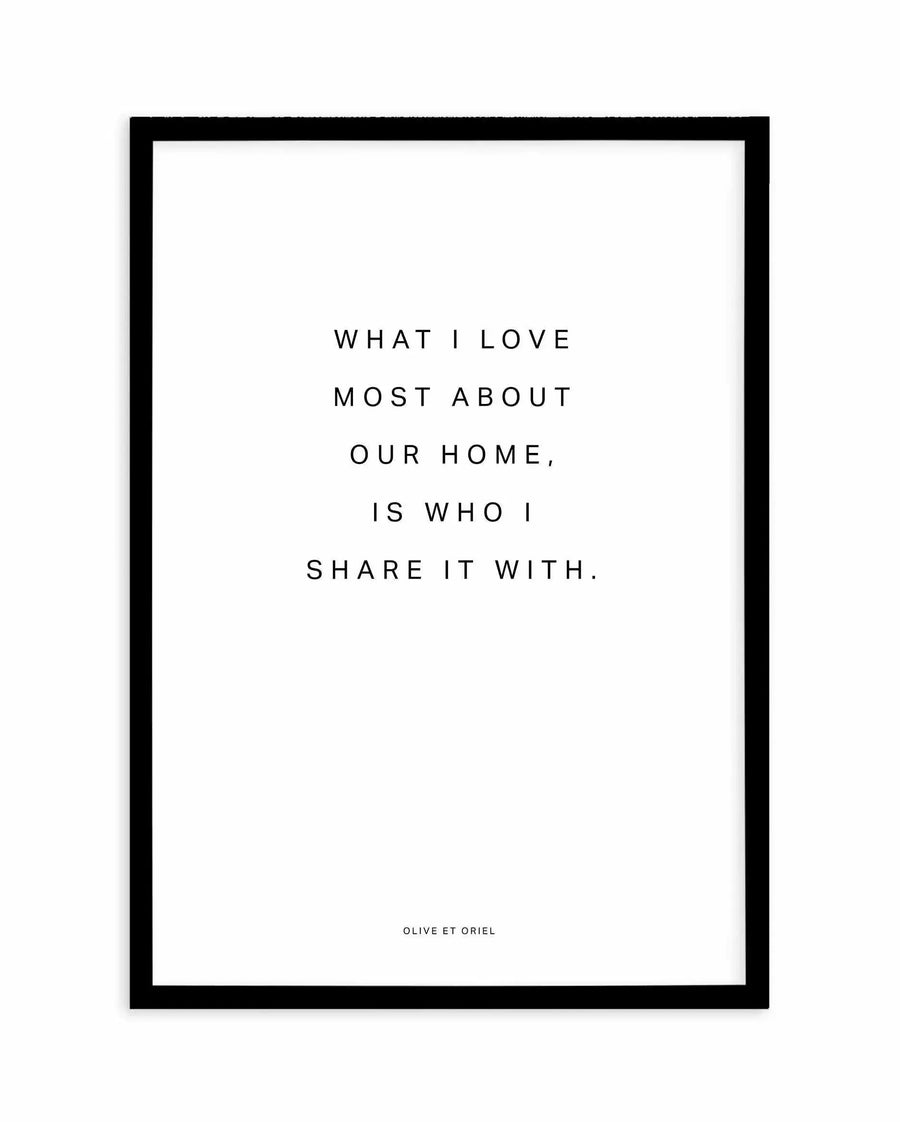 Art print with black frame featuring centered text on white background. The quote reads "WHAT I LOVE MOST ABOUT OUR HOME, IS WHO I SHARE IT WITH." in black serif font, evenly spaced across the print. Small attribution text "OLIVE ET ORIEL" appears at bottom center. The print has a clean, minimalist aesthetic with generous white space around the text. Black wooden frame with white border completes the presentation.