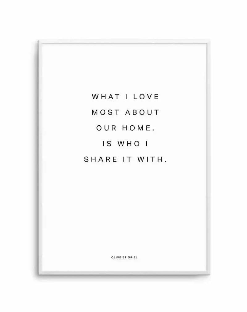 Framed art print with light gray frame displaying centered text in a simple sans-serif font reading "WHAT I LOVE MOST ABOUT OUR HOME, IS WHO I SHARE IT WITH." on a white background with white border. Small text attribution at bottom. Minimalist typography design with black text on white, clean and modern aesthetic.
