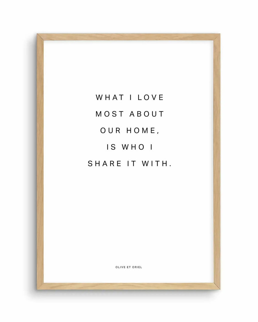 Framed art print with light natural wood frame displaying centered black text on white background that reads "WHAT I LOVE MOST ABOUT OUR HOME, IS WHO I SHARE IT WITH." Small text at bottom reads "OLIVE ET ORIEL". Minimalist typography design with clean, spaced lettering in sans-serif font.