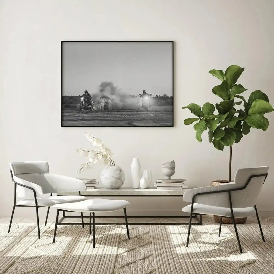 Lifestyle shot of a minimalist living room featuring a large framed black and white art print displayed above a white console table. The print is a photograph titled "Outlawed at Sundown" and depicts two motorcyclists on a dusty, barren landscape, with one creating a large cloud of dust behind them. The print is in a black frame with a white border. The room includes two light grey armchairs with black metal frames, a matching ottoman, a large green fiddle-leaf fig plant, and a textured cream rug.