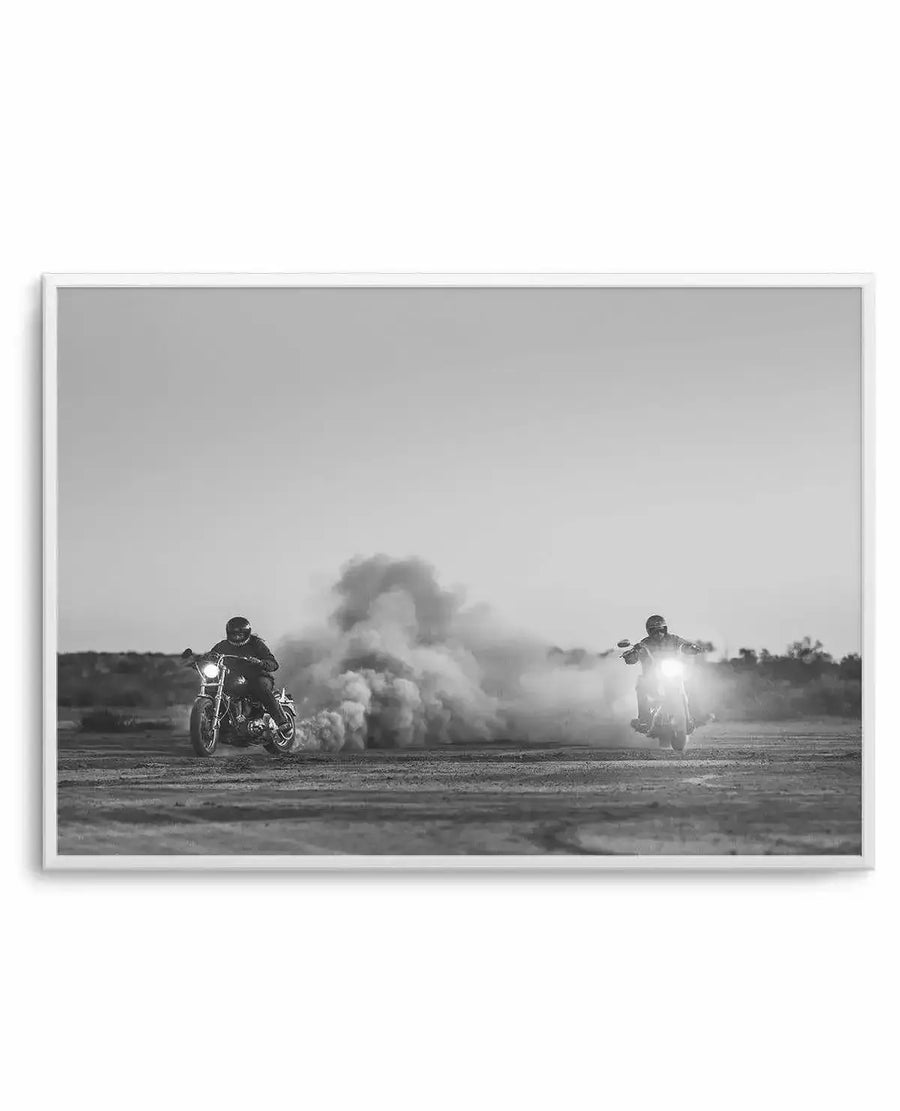 Black and white framed art print titled "Outlawed at Sundown" by Tim Harris. The photograph depicts two motorcyclists on a flat desert road with headlights illuminated, positioned on either side of a large dust cloud billowing between them. The landscape is barren and expansive under an overcast sky. The image has a dramatic, cinematic quality with high contrast monochromatic tones. White wooden frame with white border surrounding the artwork.