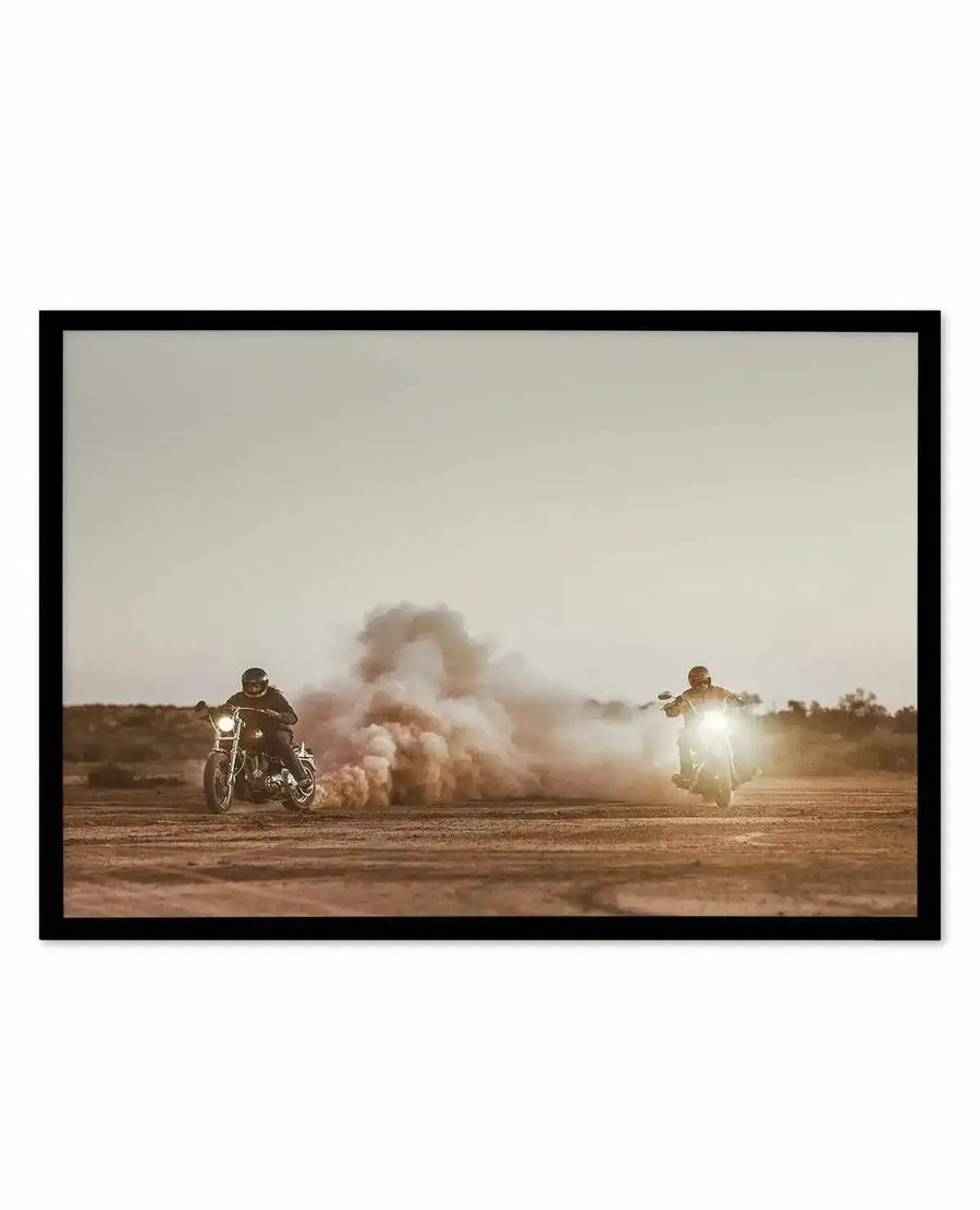 "Outlawed at Sundown" art print featuring two motorcyclists riding on a dusty desert road with warm golden sunlight, a large dust cloud billowing between them against a hazy sky, rendered in warm sepia and tan tones with a cinematic quality, displayed in a black frame with white border.