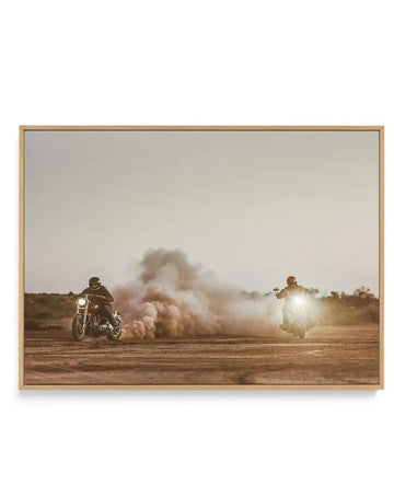 Framed canvas art print featuring a sepia-toned photograph of two motorcyclists kicking up dust in a desert landscape, with a natural oak frame. The rider on the left is closer, creating a large cloud of dust, while the rider on the right approaches with bright headlights, both under a pale, overcast sky.