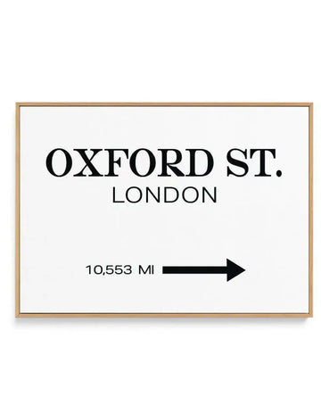 Framed canvas art print with natural wood frame displaying black text on white background. Text reads "OXFORD ST." in large serif font, with "LONDON" centered below in smaller sans-serif font. A black arrow points right next to "10,553 MI" in smaller text. The design has a minimalist, typographic style with clean lines and high contrast. White border surrounds the printed area within the wooden frame.