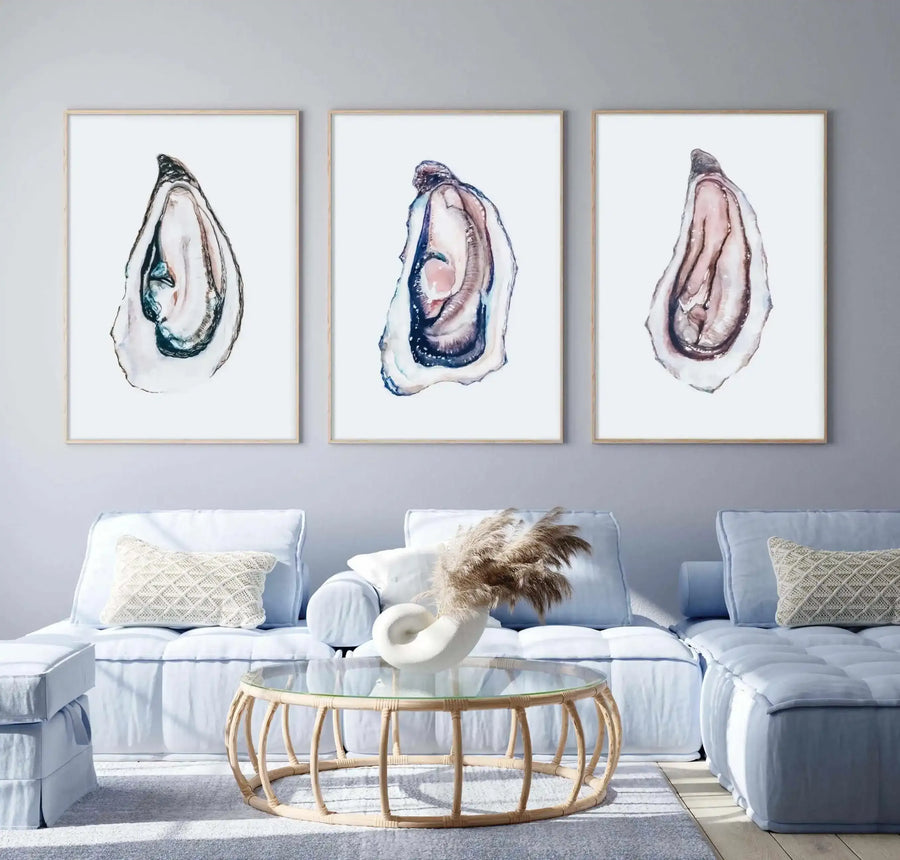 Lifestyle shot of three framed art prints displayed above a light blue sofa in a modern living room. Each print features a large-scale watercolor illustration of an oyster shell with iridescent blues, purples, and pinks on a white background, displayed in a natural oak frame with a white border. The room includes a round rattan coffee table with a glass top, a white ceramic vase with dried pampas grass, and textured cream throw pillows.