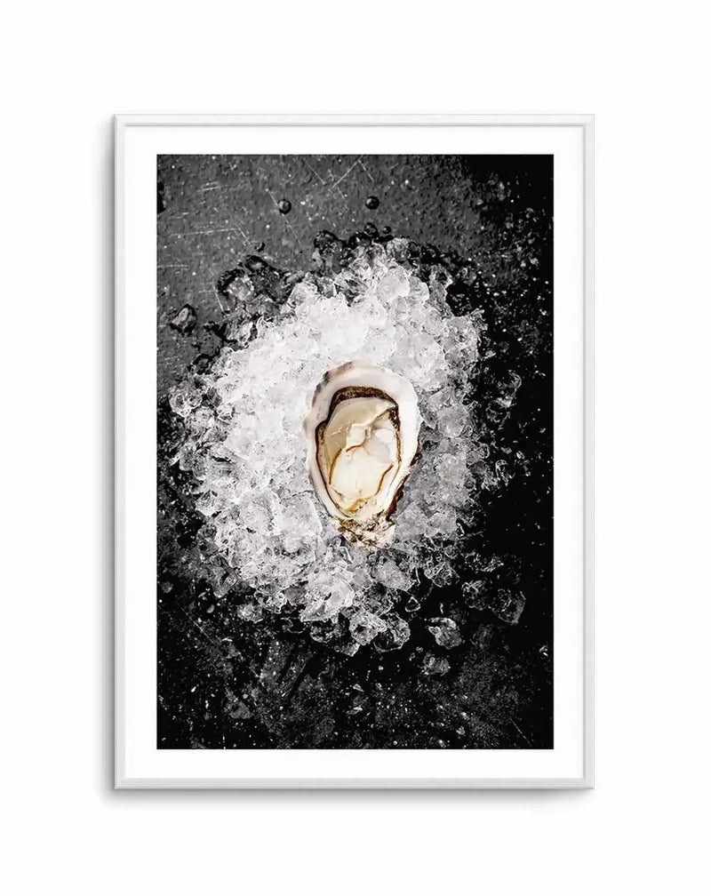Framed black and white photograph of a single oyster on a bed of crushed ice, displayed in a white frame with a white border. The oyster, with its creamy white flesh and dark edges, is centered on a dark, textured surface, possibly slate or stone, with scattered water droplets.