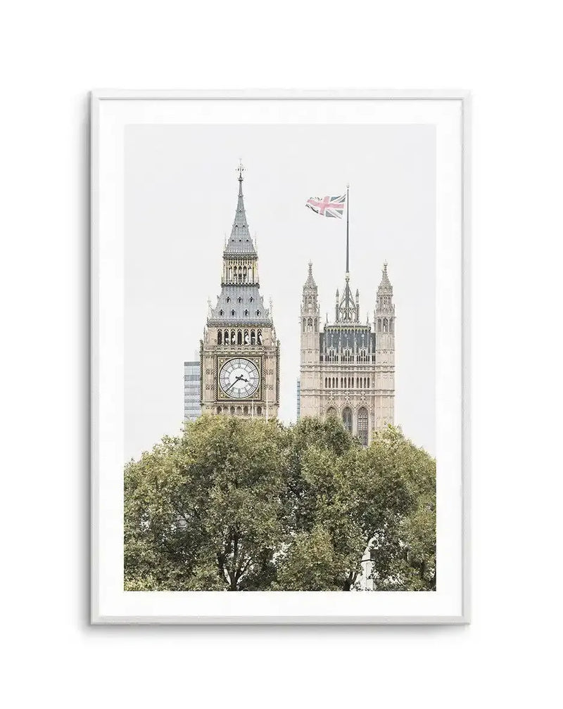 Framed photograph of the Palace of Westminster in London, England, with a white frame and white border. The composition features Big Ben and the Houses of Parliament partially obscured by lush green trees in the foreground, under a bright white sky. The Union Jack flag flies above the Houses of Parliament.