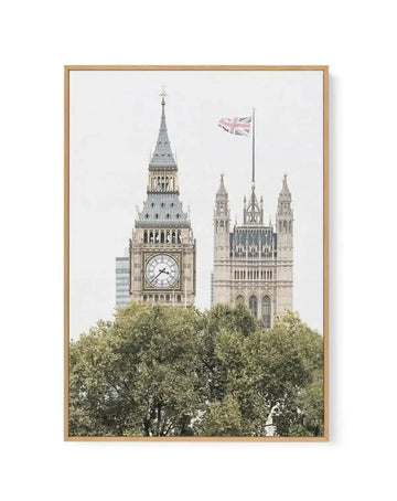 Framed canvas art print featuring a bright, high-key photograph of the Palace of Westminster in London, England. The composition shows Big Ben (Elizabeth Tower) on the left with its clock face visible, and the Houses of Parliament on the right with the Union Jack flag flying above. The lower portion of the image is filled with lush green trees, obscuring the base of the buildings. The print is displayed in a natural oak frame.