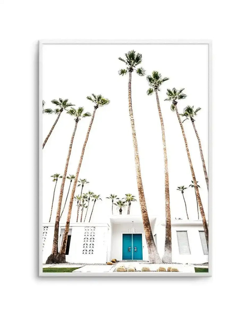Framed art print featuring a minimalist photograph of a white mid-century modern building with a turquoise-blue double door, flanked by tall palm trees with textured brown trunks and green fronds. Small cacti in terracotta pots sit at ground level. The composition is shot from a low angle against a bright white sky, creating clean vertical lines. The image has a serene, desert modernist aesthetic with muted earth tones. The print is mounted in a white frame with white border.