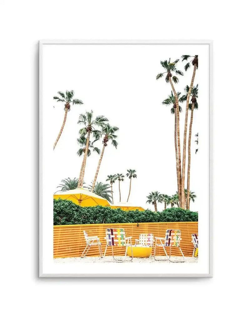 Framed art print featuring a minimalist desert scene with tall palm trees against a white sky. Yellow umbrellas and colorful striped beach chairs are arranged along an orange corrugated metal fence in the foreground, with green hedging visible behind. The composition captures a bright, sunny aesthetic typical of Palm Springs. The print is displayed in a white frame with a white border.