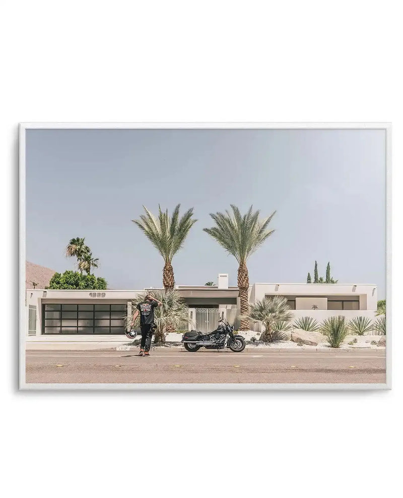 Framed photograph of a man standing next to a black motorcycle in front of a modern white house with two large palm trees. The man, dressed in black, holds a helmet and looks towards the house, which has a two-car garage with dark glass doors and the number "1389" above it. The scene is set on a sunny day with a clear, pale blue sky, and the foreground features a paved road with yellow markings.