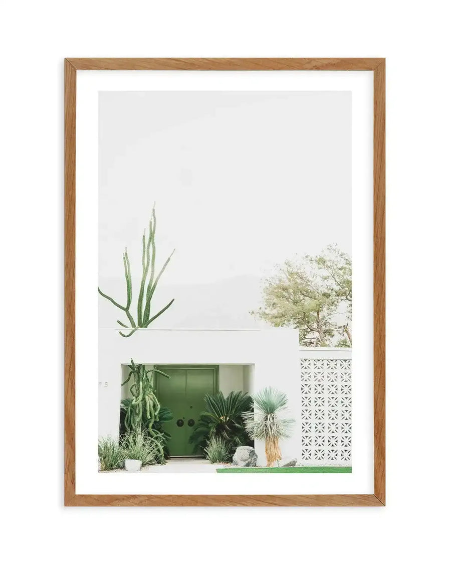 Framed art print featuring a minimalist architectural photograph of a mid-century modern desert home. The image shows a white-walled facade with a vibrant sage green garage door, surrounded by desert plants including tall agave, fan palms, and ornamental grasses in cream and green tones. A decorative white geometric screen is visible on the right, with a pale tree visible above. The photograph is mounted with a white border in a natural wood frame with visible grain texture.