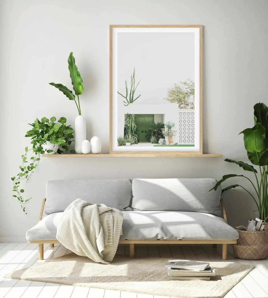 Lifestyle shot of a minimalist living room featuring a framed art print above a light grey sofa. The print is a photograph of a white building with a vibrant green door, surrounded by various green plants and cacti under a bright sky, framed in natural oak with a white border. The room includes a light wood shelf with potted plants and white vases, a cream throw blanket on the sofa, and a woven basket with magazines on the floor.