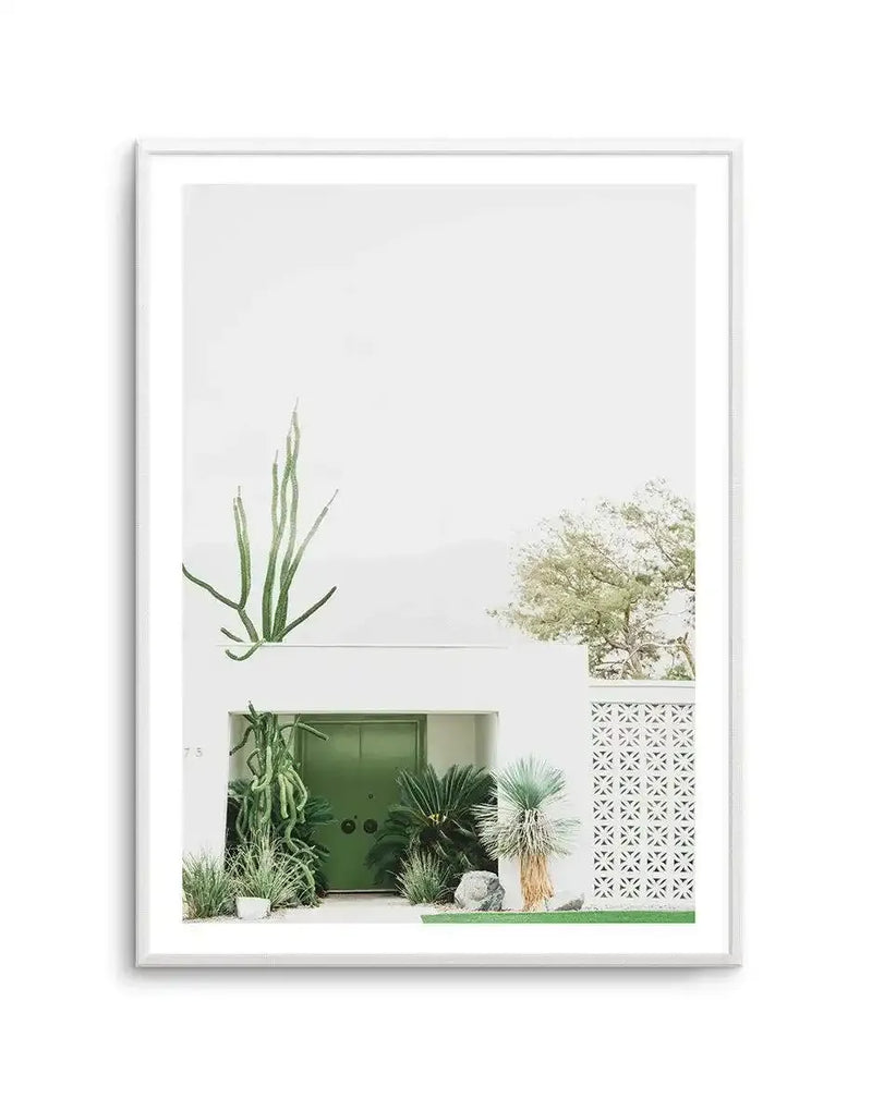 Framed photography art print featuring a minimalist white building with a bright green door and desert landscaping, displayed in a white frame with a white border. The composition shows a tall, slender cactus on the left, a vibrant green door surrounded by various succulents and palm-like plants, and a white decorative screen on the right, all under a bright, clear sky.