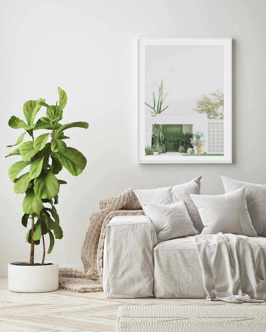 Lifestyle shot of a minimalist living room featuring a framed art print above a light grey sofa. The print is a photograph of a modern white building with a vibrant green door, surrounded by various cacti and desert plants, including a tall, multi-stemmed cactus hanging from above. The room has light wooden flooring, a large fiddle leaf fig plant in a white pot, a chunky knit cream throw, and several light grey cushions on the sofa.