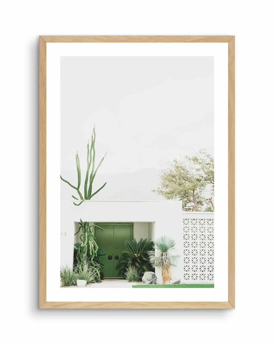 "Framed photography art print of a minimalist white building with a bright green double door, surrounded by desert plants and cacti, displayed in a natural oak frame with white border. The composition features a tall, slender cactus on the left, a white breeze block wall on the right, and a light-colored tree in the background against a bright, overcast sky."