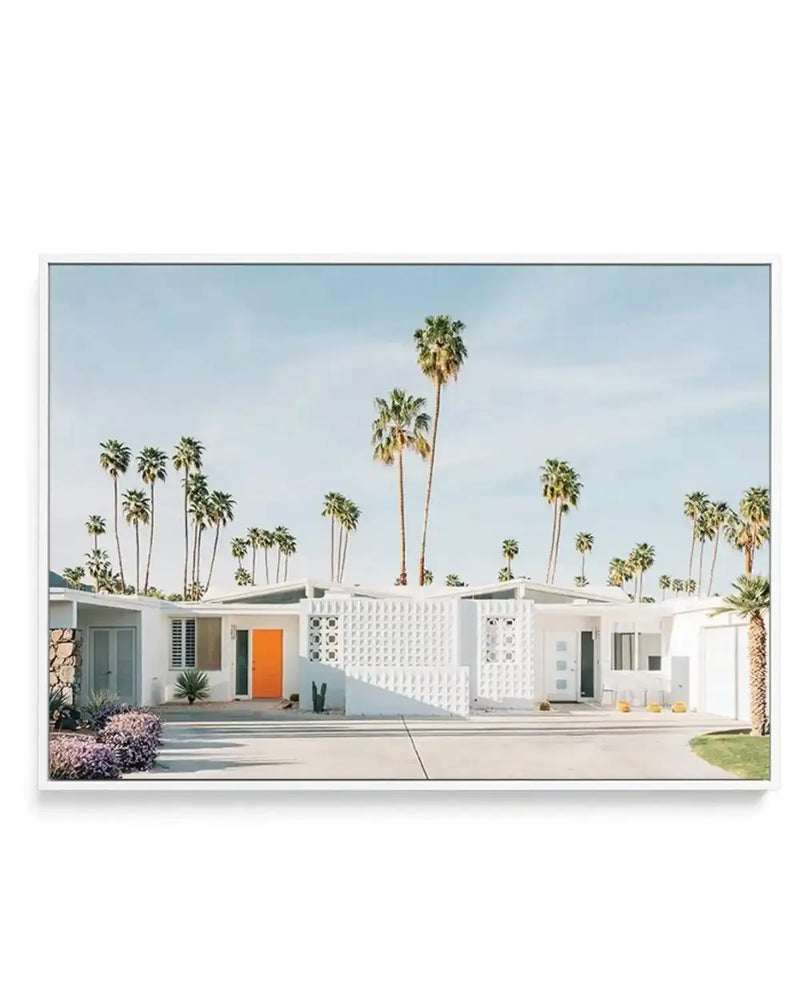 Framed canvas art print depicting a mid-century modern white single-story house with an orange front door and decorative white lattice panel, set against a row of tall palm trees under a light blue sky. The house features a manicured front lawn with purple flowering shrubs and desert landscaping. The canvas is mounted in a slim gray frame with a white border.